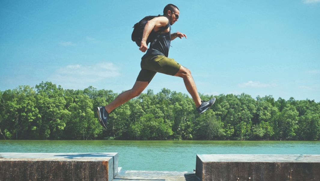 Man Leaping On Concrete Surface Near Body Of Water And Forest At The Distance During Day